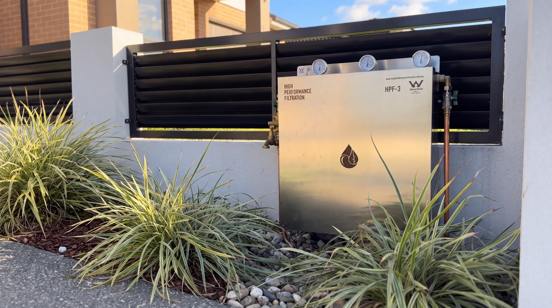A whole house water filtration system installed at the front fence of a modern Sydney home, pressure gauges on top and a garden bed around it
