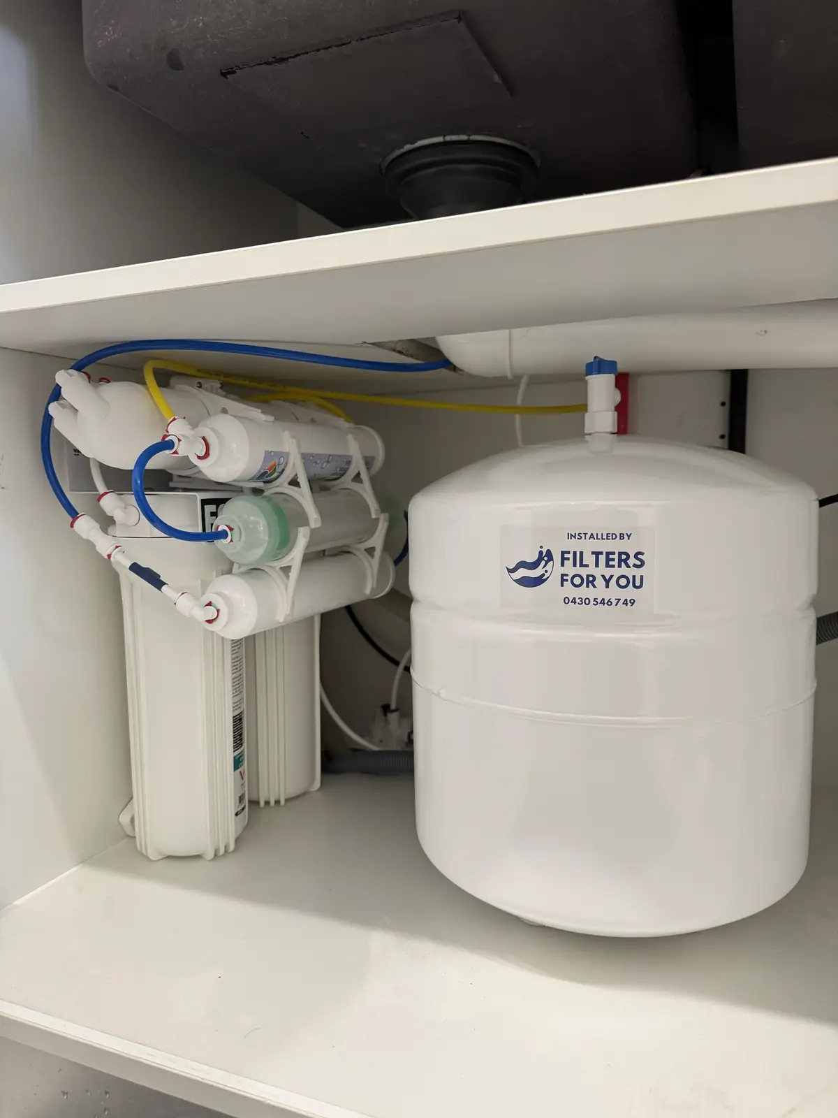 A reverse osmosis system and storage tank installed under a kitchen sink, with an Installed by Filters For You sticker on the tank